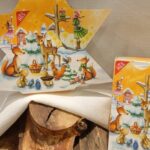 Adventskalender-Tee in Sternform und normaler rechteckiger Verpackung dekoriert auf einem weißen Tuch mit Holzstämmen.