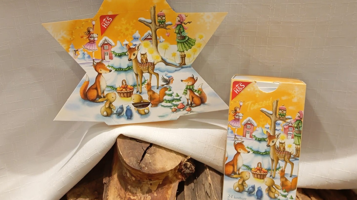 Adventskalender-Tee in Sternform und normaler rechteckiger Verpackung dekoriert auf einem weißen Tuch mit Holzstämmen.